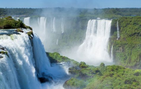 Panoramic view of Iguazu Falls, featuring multiple waterfalls cascading over lush green cliffs, mist rising, and a vibrant rainbow arching at the base.