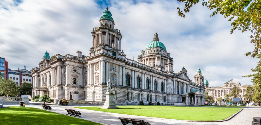 Historic stone building with green domes, ornate columns, and statues, set in a park with lush grass and trees. The sky is partly cloudy, creating a serene atmosphere.