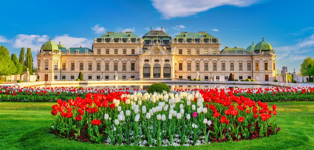 A grand Baroque palace with a green roof is framed by vibrant red and white tulips in lush gardens under a bright blue sky with wispy clouds.