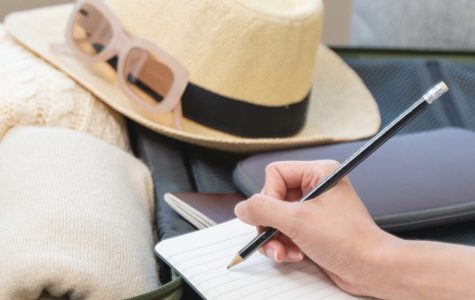 Person writing in a notebook next to an open suitcase filled with clothes, headphones, a camera, hat, and sunglasses, conveying a travel preparation vibe.