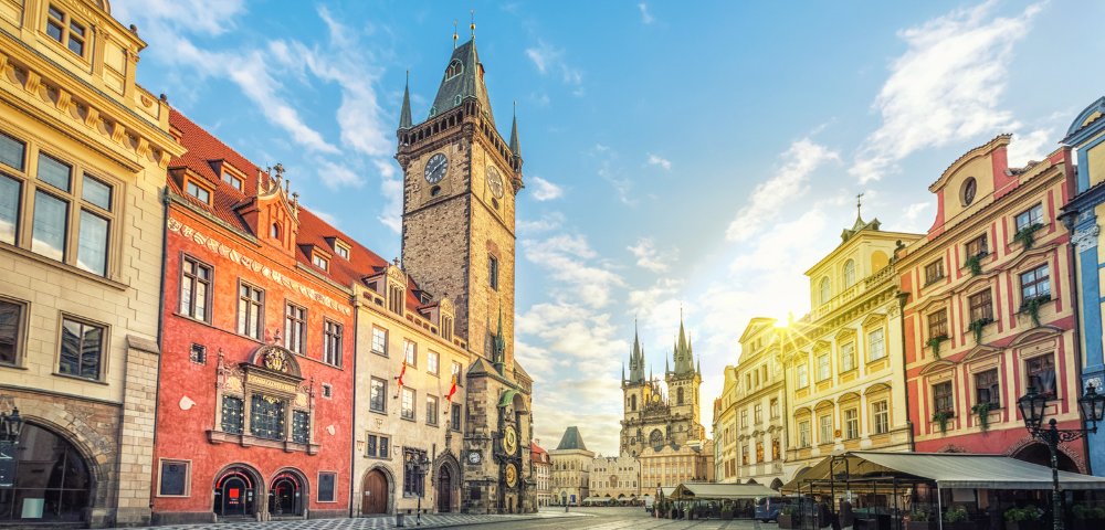 A sunlit European square with historic buildings, including a clock tower and church, under a partly cloudy sky. The scene is serene and picturesque.