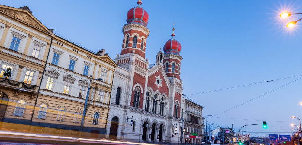 Historic building with red domes and intricate architecture at dusk, illuminated by streetlights. Blurred car lights suggest bustling street activity.
