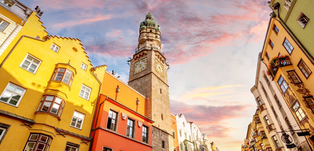 Colorful buildings with a medieval clock tower under a vibrant sunset sky. The scene conveys a warm, historical ambiance with pastel hues.