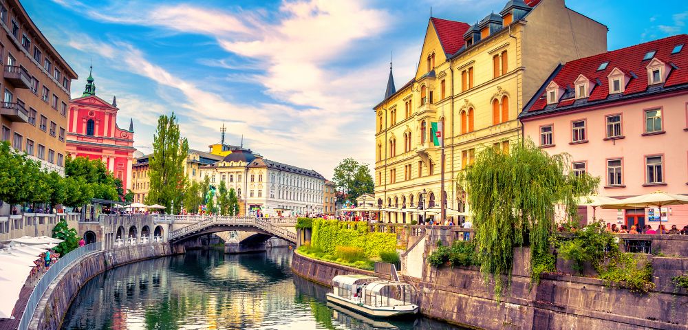 A picturesque European canal scene features colorful historic buildings, a stone bridge, and lush greenery under a vibrant blue sky with wispy clouds.