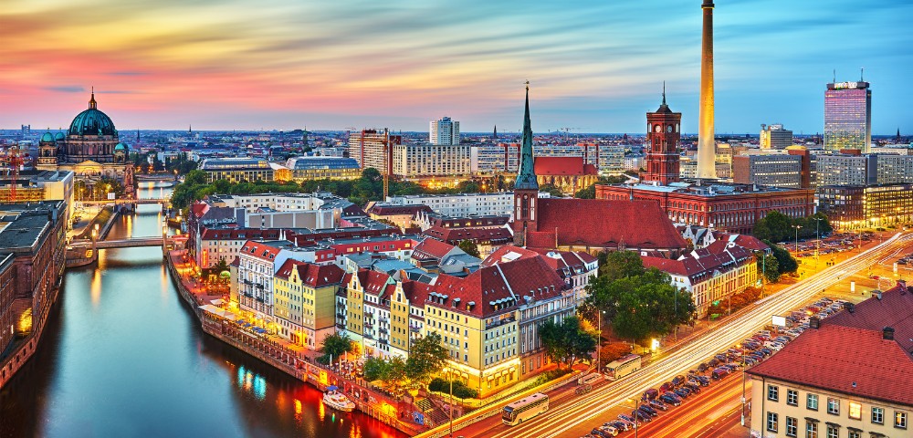 A vibrant cityscape of Berlin at sunset, featuring the Spree River, historic buildings with red roofs, and a colorful sky, creating a lively and serene atmosphere.
