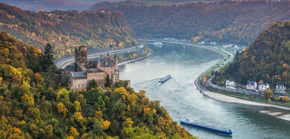 Aerial view of a historic castle amid vibrant autumn trees overlooking a winding river with boats. The scene is serene and picturesque.