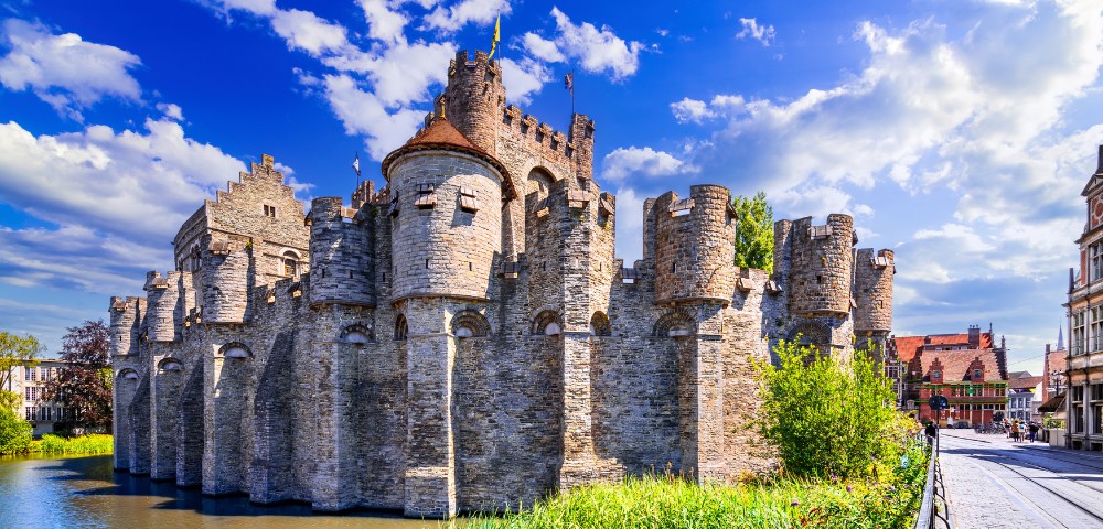 Majestic medieval castle with round towers and a crenellated wall, surrounded by a moat on a sunny day. Blue sky and fluffy clouds above.
