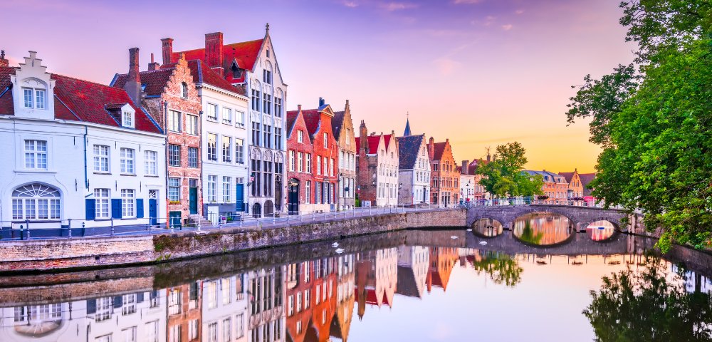 A picturesque canal in Bruges at sunset, reflecting colorful, historic European buildings and a stone bridge in calm water, surrounded by lush trees.