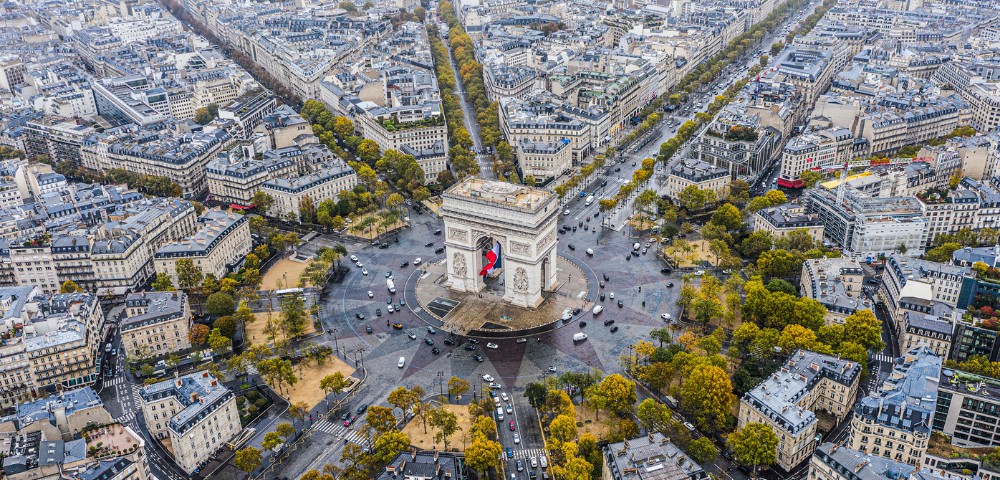 Aerial view of the Arc de Triomphe in Paris, surrounded by busy streets and autumn-colored trees, radiating a sense of vibrant city life and grandeur.