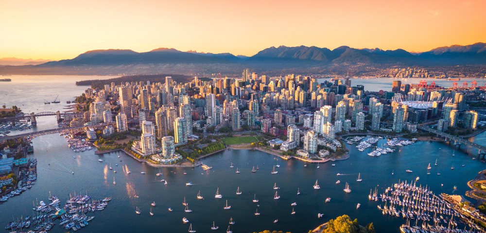Aerial view of a vibrant cityscape at sunset with numerous skyscrapers surrounded by water and boats. Mountains in the background enhance the serene atmosphere.