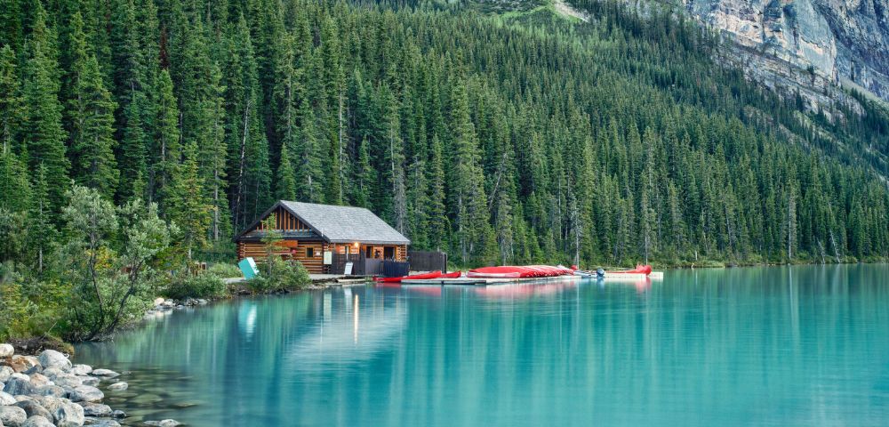 A serene lakeside scene with a wooden cabin beside a teal lake, surrounded by dense evergreen forest and mountains. Canoes are lined up on the dock.