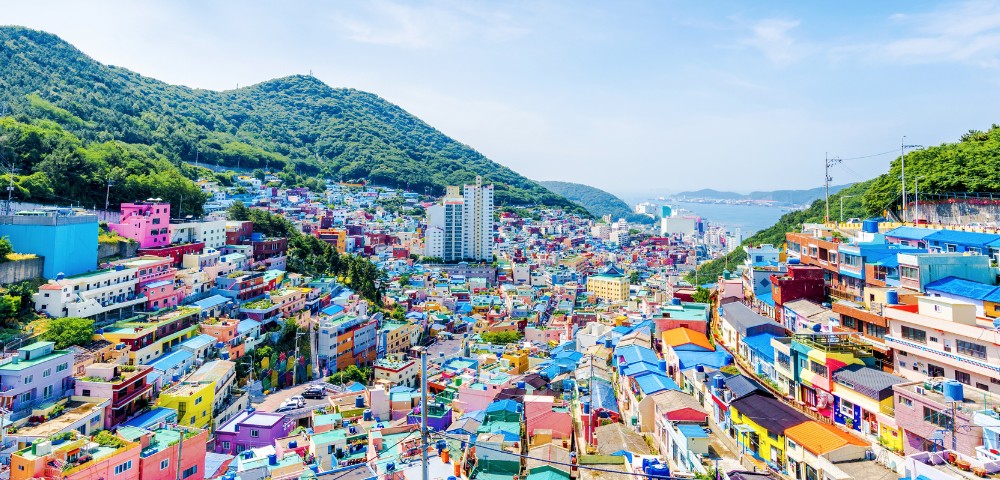 Colorful hillside village with vibrant houses, nestled against lush green mountains. The scene conveys a lively, cheerful atmosphere under a clear blue sky.
