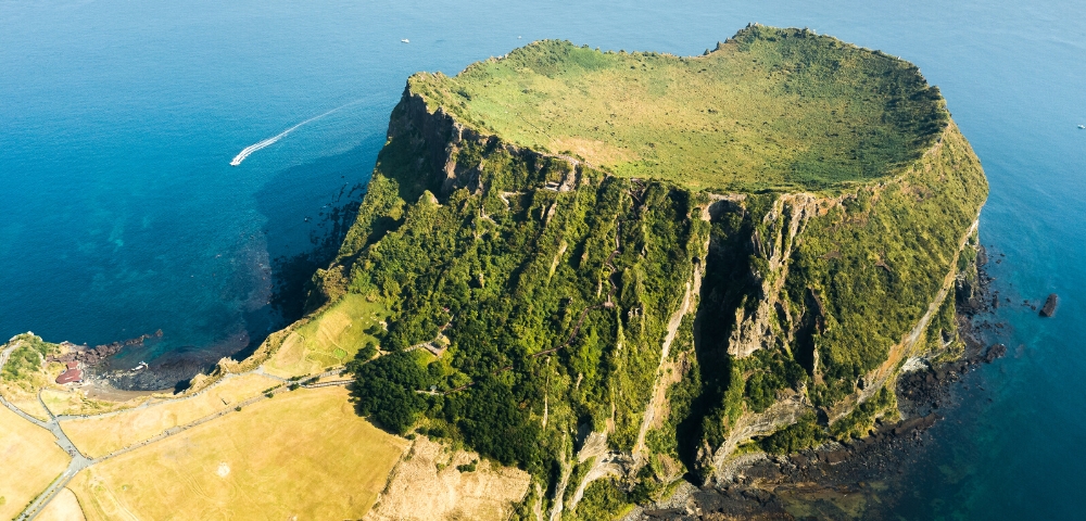 Aerial view of a volcanic crater island surrounded by the ocean. The lush green top contrasts with steep rocky cliffs. A boat leaves a trail in the blue water.