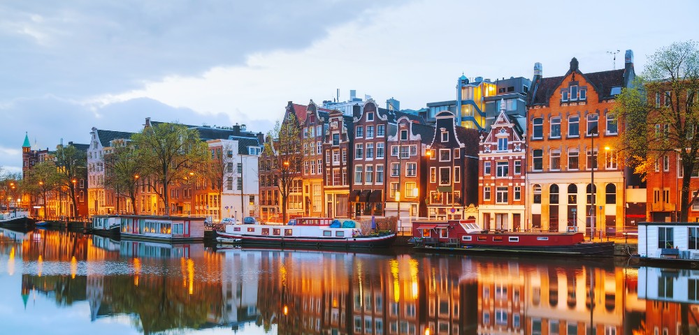 Scenic view of Amsterdam's iconic, illuminated canal houses at dusk, reflecting on the tranquil water, lined with houseboats and trees.