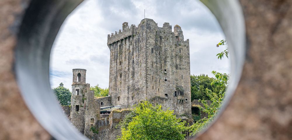 Stone castle with a tall tower framed by a circular opening. Surrounded by greenery and set against a cloudy sky, evoking a historic and mysterious feel.