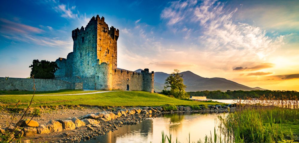A medieval stone castle stands by a tranquil river, surrounded by lush greenery. The sky glows with a colorful sunset, creating a serene and majestic atmosphere.