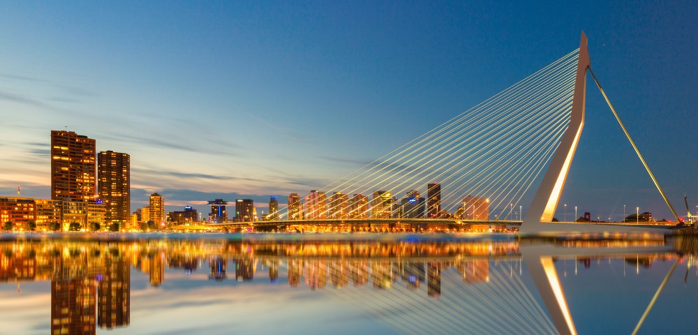 Night view of a brightly lit cable-stayed bridge with twinkling city lights reflected on calm water, set against a clear blue sky, creating a serene mood.