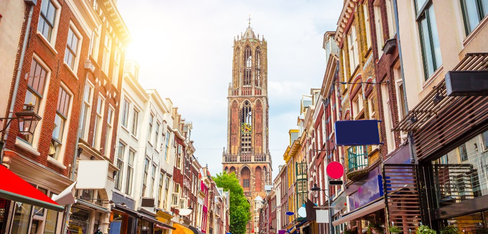 A sunny street view leads to a tall, ornate tower, surrounded by charming, colorful buildings with awnings. The scene feels lively and historic.