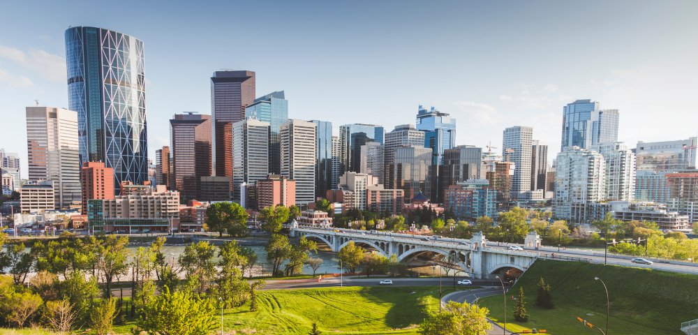 A bright cityscape of Calgary with modern skyscrapers under a clear sky. A lush green park and bridge with cars are in the foreground.