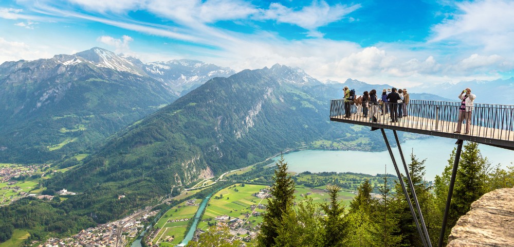A scenic mountainside view with a group of people standing on a viewing platform overlooking lush green valleys, a lake, and distant peaks under a bright blue sky.