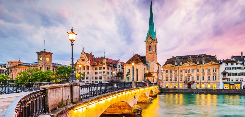Scenic view of Zurich's old town at sunset, featuring a bridge over the Limmat River, historic buildings, a prominent spire, and warm street lighting.