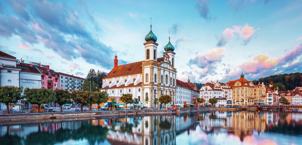 A picturesque scene of a baroque-style church with twin green-domed towers at sunset, reflecting in calm river waters, surrounded by quaint buildings.