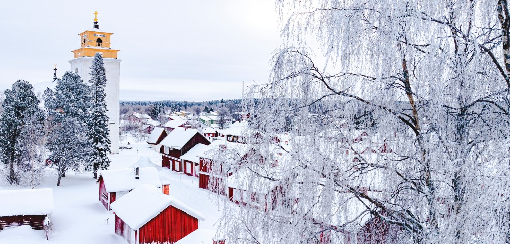 Winter scene of a village covered in snow, with red wooden houses and a yellow-topped tower. Frost-covered trees evoke a serene, cold atmosphere.