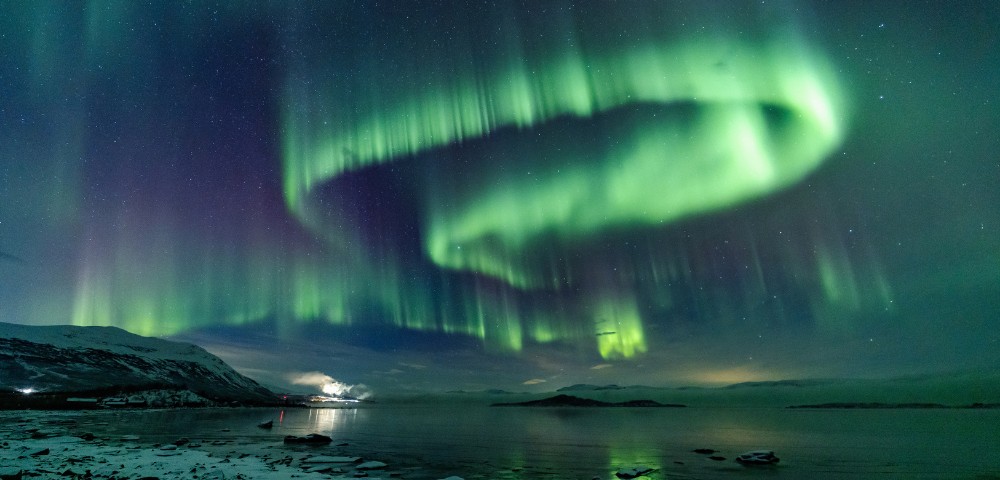 Vibrant green and purple aurora borealis lights up a starry night sky over a calm arctic landscape with snowy mountains and a reflective lake.
