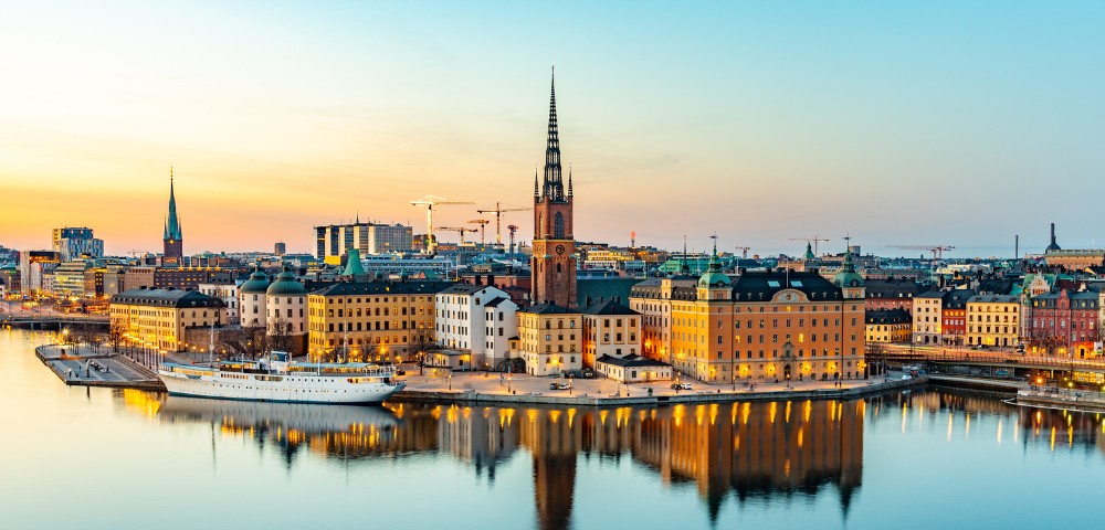 Panoramic view of Stockholm at sunset, featuring historic buildings and a church spire reflected in serene waters. Warm, tranquil hues dominate the scene.