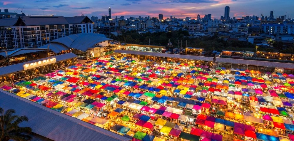 Aerial view of a vibrant night market with rows of colorful tents illuminated against a city skyline at dusk. Atmosphere is lively and bustling.