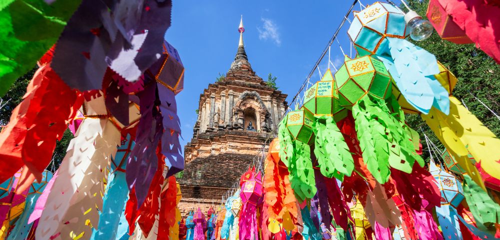 Colorful paper lanterns hang vibrantly in front of a historic, intricately carved brick temple tower under a clear blue sky, creating a festive and lively atmosphere.