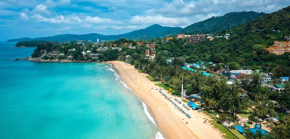 Aerial view of a serene beach with turquoise waters and golden sand, bordered by lush green hills. Buildings are visible among the trees. Bright and calm.