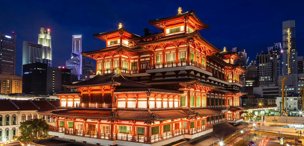 Night view of the Buddha Tooth Relic Temple in Singapore, illuminated in vibrant red and gold lights, with a backdrop of modern skyscrapers.