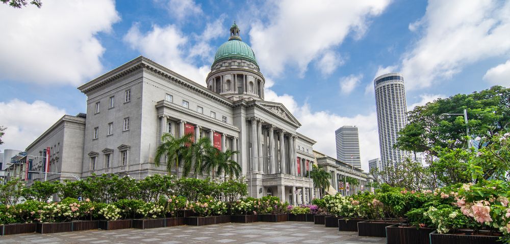 Historic neoclassical building with a green dome, surrounded by lush plants and modern skyscrapers under a partly cloudy sky.
