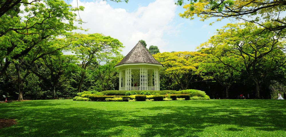 White gazebo surrounded by lush green trees and manicured lawn under a partly cloudy blue sky in a park.