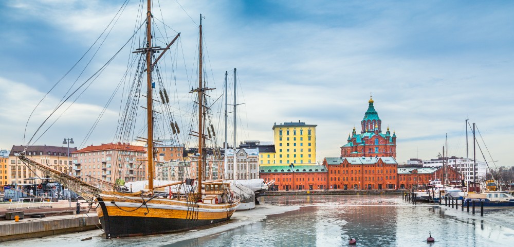Sailboats docked along a calm harbor with colorful historic buildings and a domed church in the background under a cloudy sky.