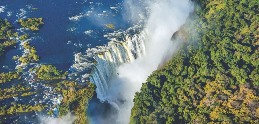 Aerial view of a wide waterfall surrounded by dense green forest and flowing blue river water.