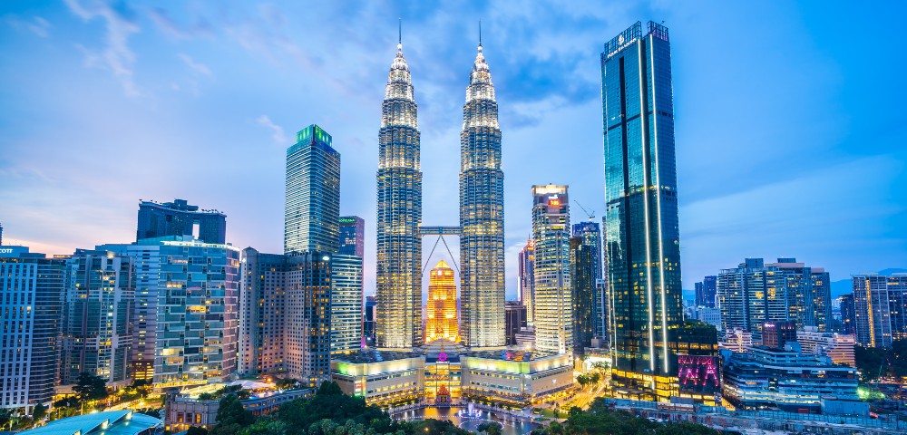Cityscape at dusk featuring illuminated twin skyscrapers, surrounded by modern buildings. The sky is a soft blue, enhancing the vibrant urban atmosphere.