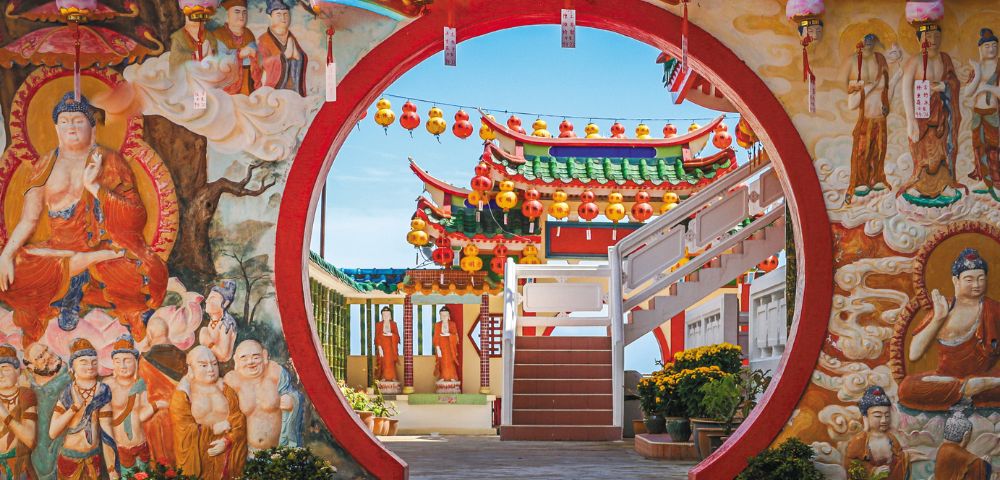 Circular red archway frames a vibrant Chinese temple courtyard with colorful lanterns and intricate wall murals of deities under a clear blue sky.