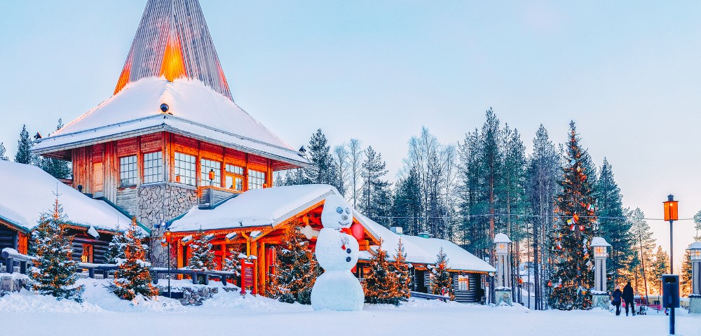 Snowy winter scene at a cozy wooden lodge with a large snowman in front. Surrounded by snow-covered pine trees under a clear blue sky, evoking a festive, tranquil atmosphere.