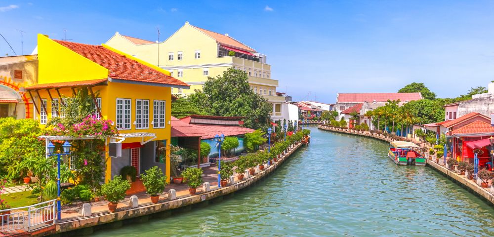 A vibrant canal scene with colorful houses lining the waterway. A bright yellow house is prominent, while greenery and flowers add a cheerful atmosphere.