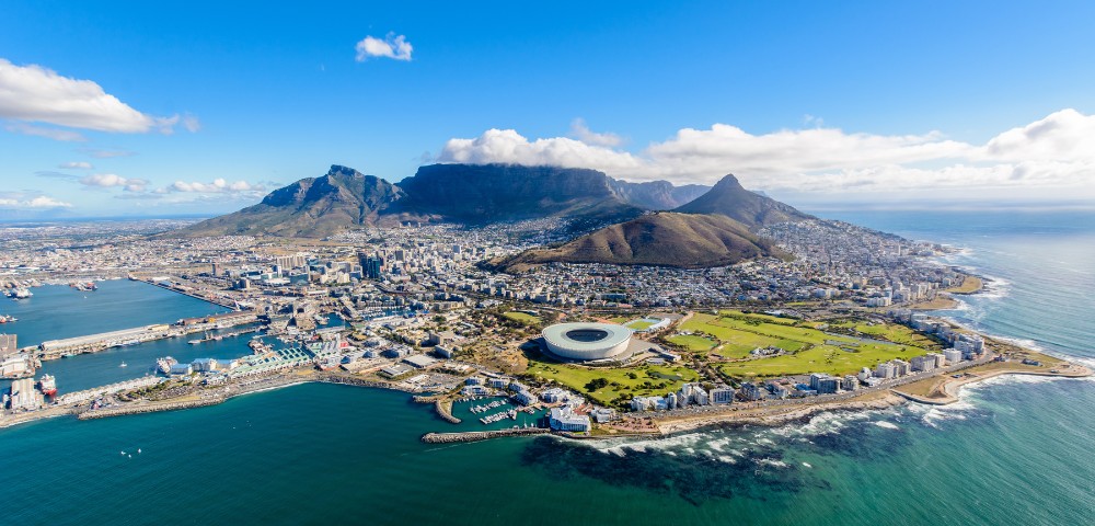 Aerial view of Cape Town, showing Table Mountain in the background, a large stadium near the coast, lush greenery, and the vibrant urban landscape.