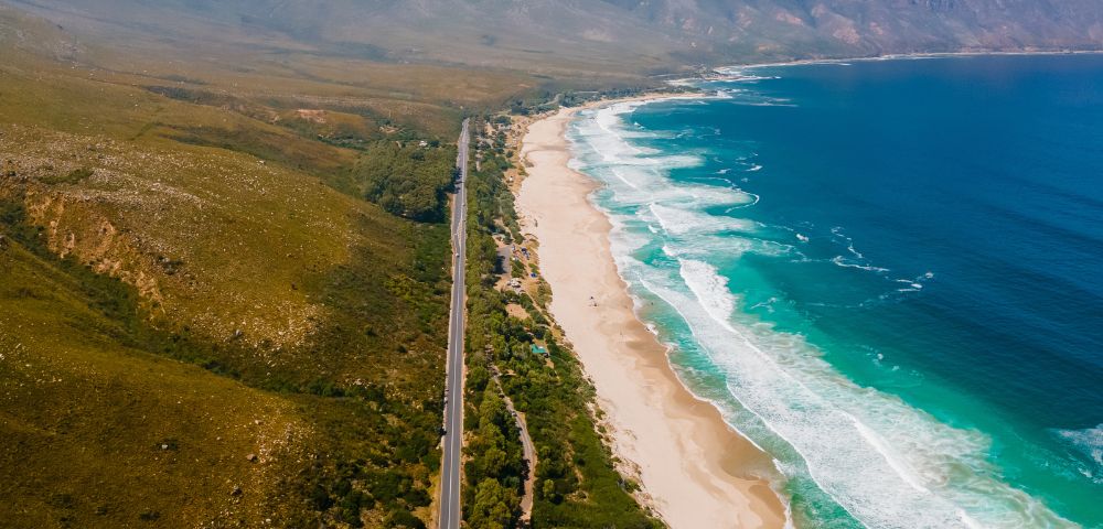 Aerial view of a scenic coastal road beside a long beach with turquoise ocean waves. Green hills stretch parallel, creating a serene, natural landscape.
