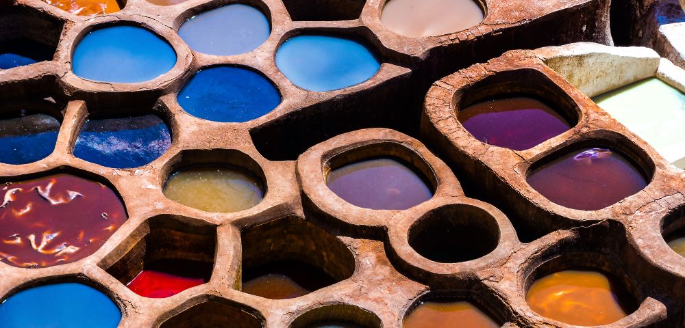 Aerial view of vibrant, round dye pits in a tannery. Each pit contains liquid of various colors like blue, red, and orange. The scene is vivid and artistic.