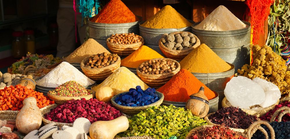 Vibrant market scene with bowls and baskets displaying colorful spices and dried flowers. Warm sunlight highlights rich reds, yellows, and blues. Energetic and lively.