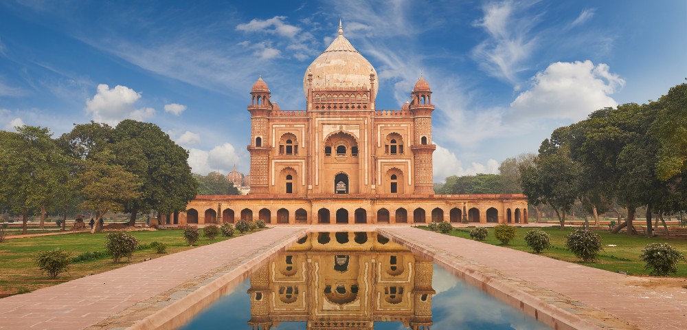 Historic red sandstone mausoleum with large dome and arched facade, reflected in a tranquil pond. Flanked by lush green trees under a clear blue sky.