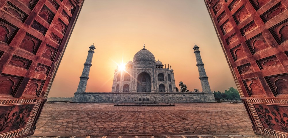 The Taj Mahal viewed through an arched doorway at sunrise, with the sun glowing behind the domed structure, against a warm, serene sky.