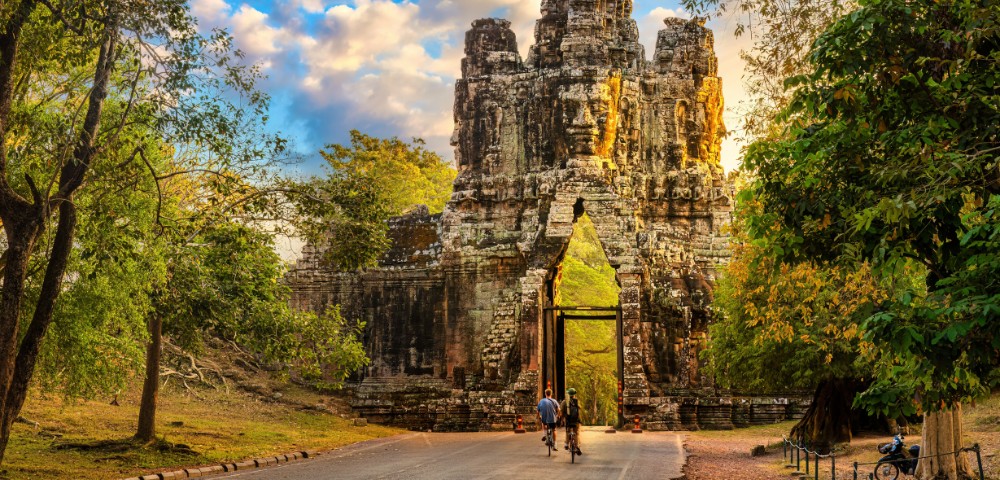 A serene scene of two cyclists approaching an ancient stone gateway surrounded by lush trees, with golden sunlight casting a warm glow over the path.