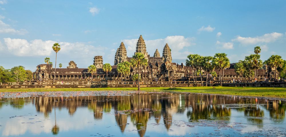 The image shows Angkor Wat, a majestic stone temple complex in Cambodia, with tall spires and palm trees reflecting in a serene pond under a blue sky.