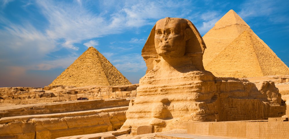 The Great Sphinx of Giza with the pyramids in the background under a blue sky. The ancient monuments convey a sense of grandeur and history.